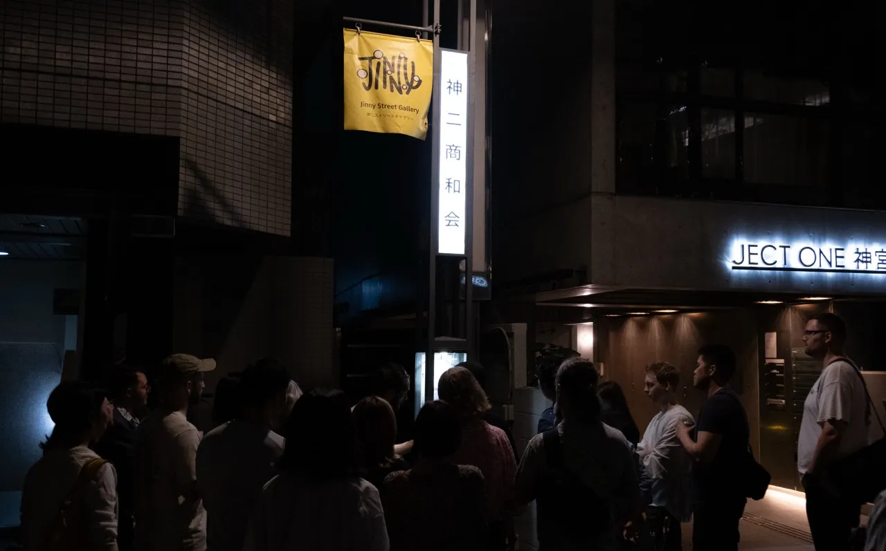 Visitors stand in the dark under a streetlamp flying the yellow Jinny Street Gallery banner on opening night